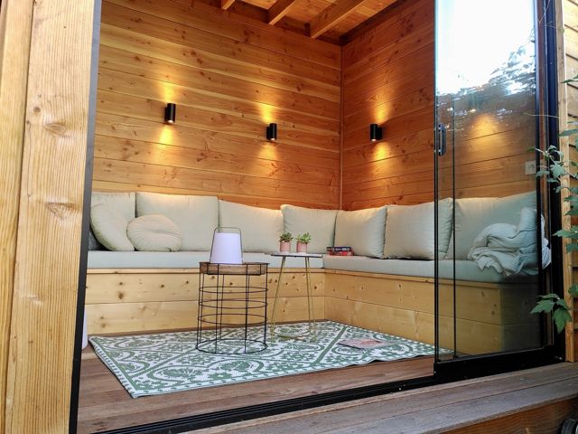 Interieur van een tuinkamer met loungehoek, kussens en wandverlichting achter een glazen schuifwand